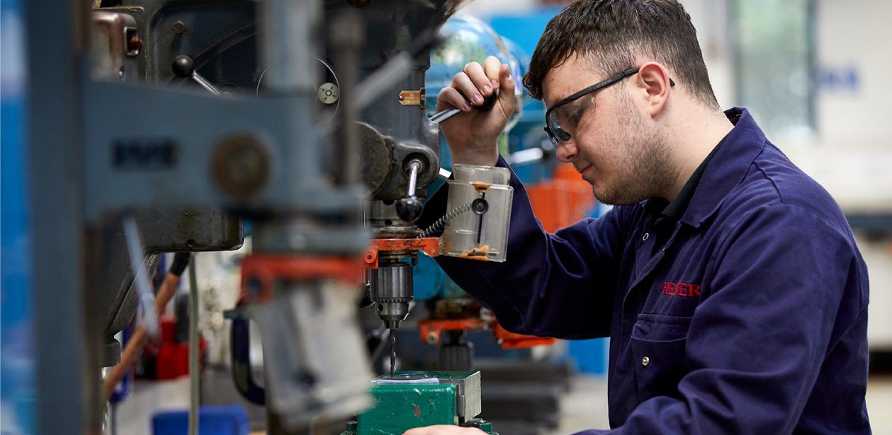 A person wearing safety glasses and a blue work uniform focuses on operating a drill press in an industrial workshop.