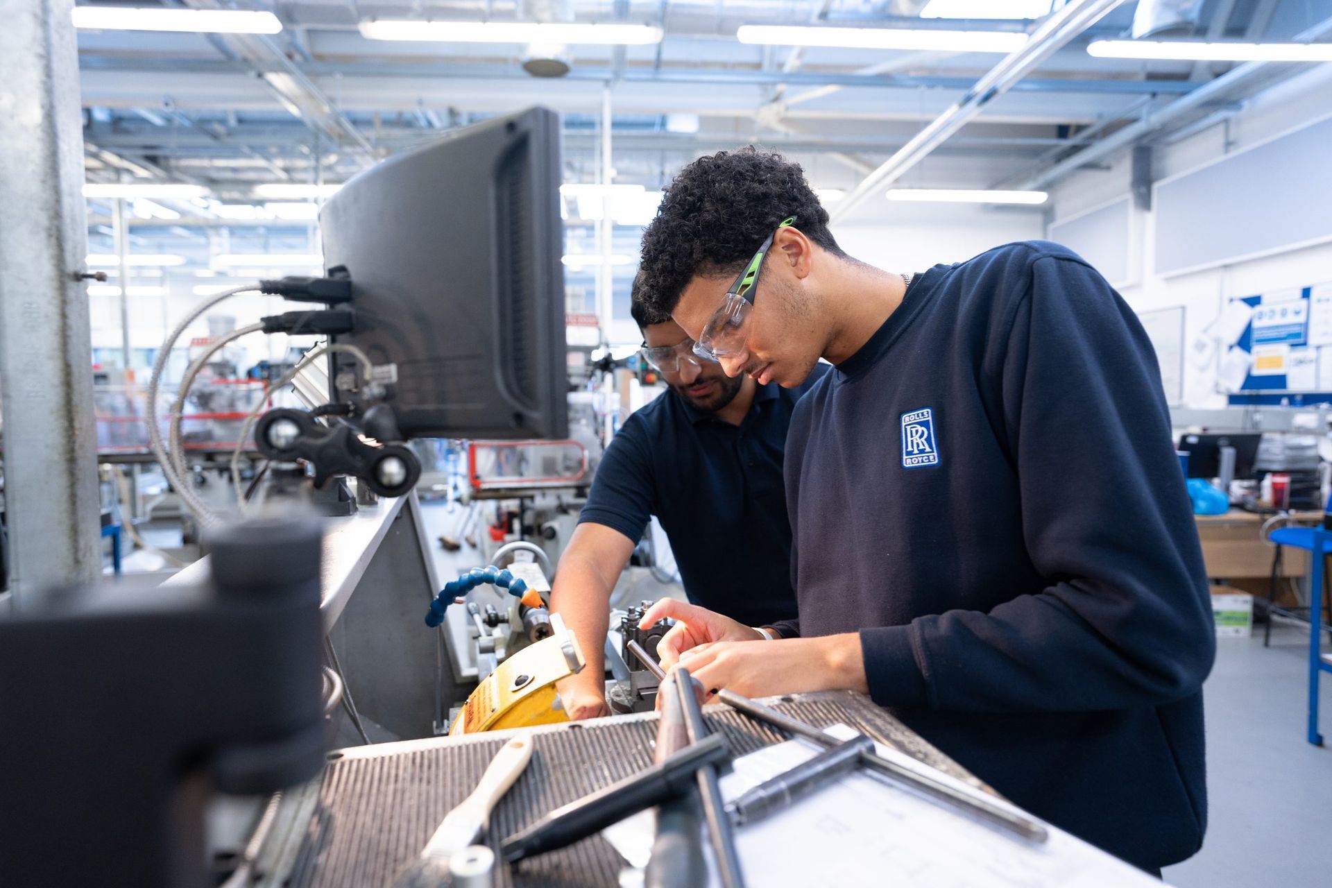 Two people wearing safety glasses work together on a project at a computer station in an industrial workshop.
