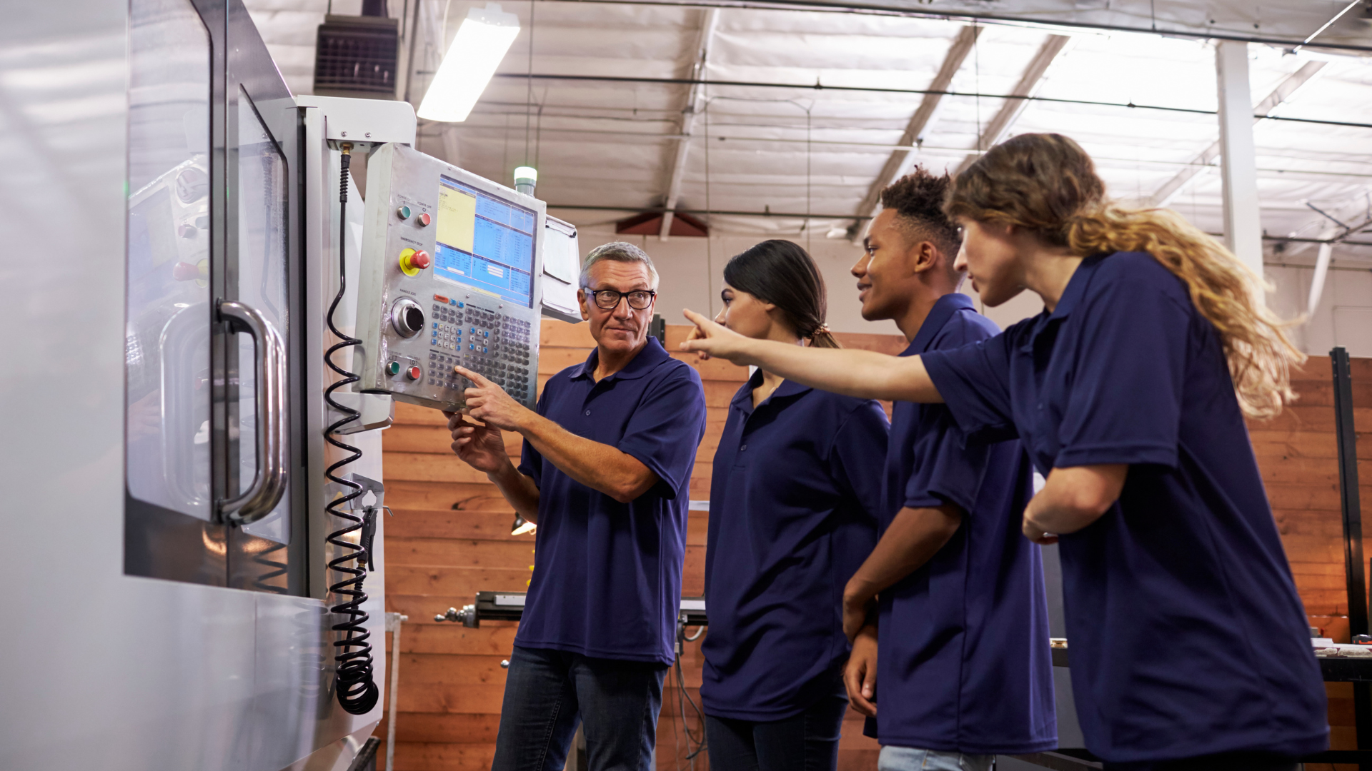 An instructor explains the control panel of a manufacturing machine to three students in a workshop setting.