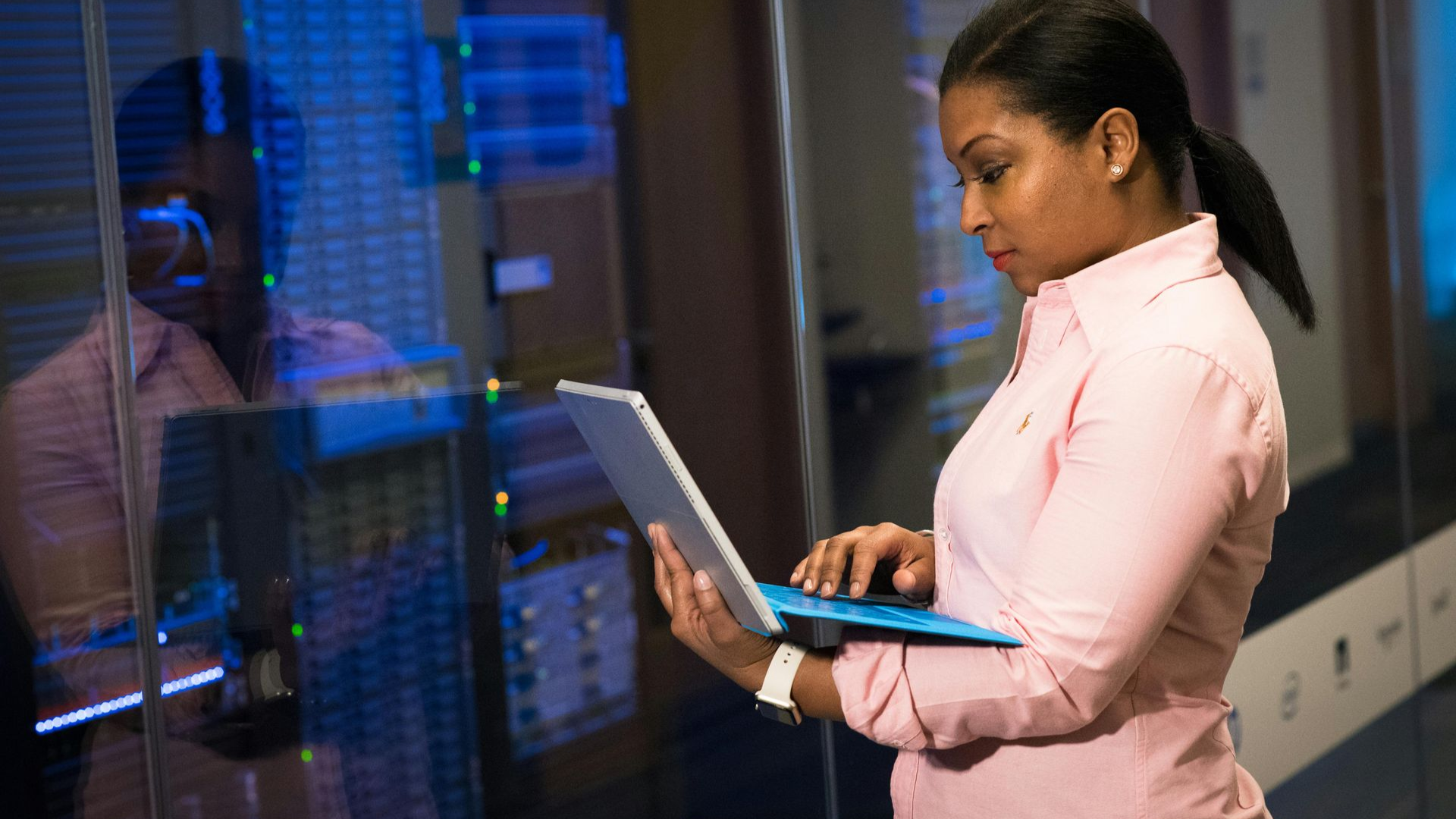 A professional checks a laptop while standing in a server room with blue-lit equipment racks in the background.