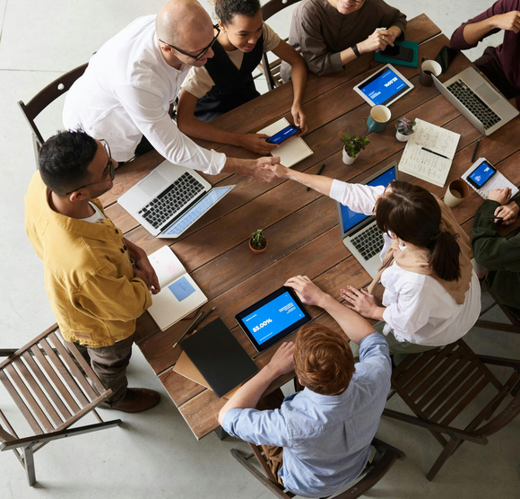 A diverse group works around a wooden table with laptops, notebooks, and tablets, while two people shake hands in the center.
