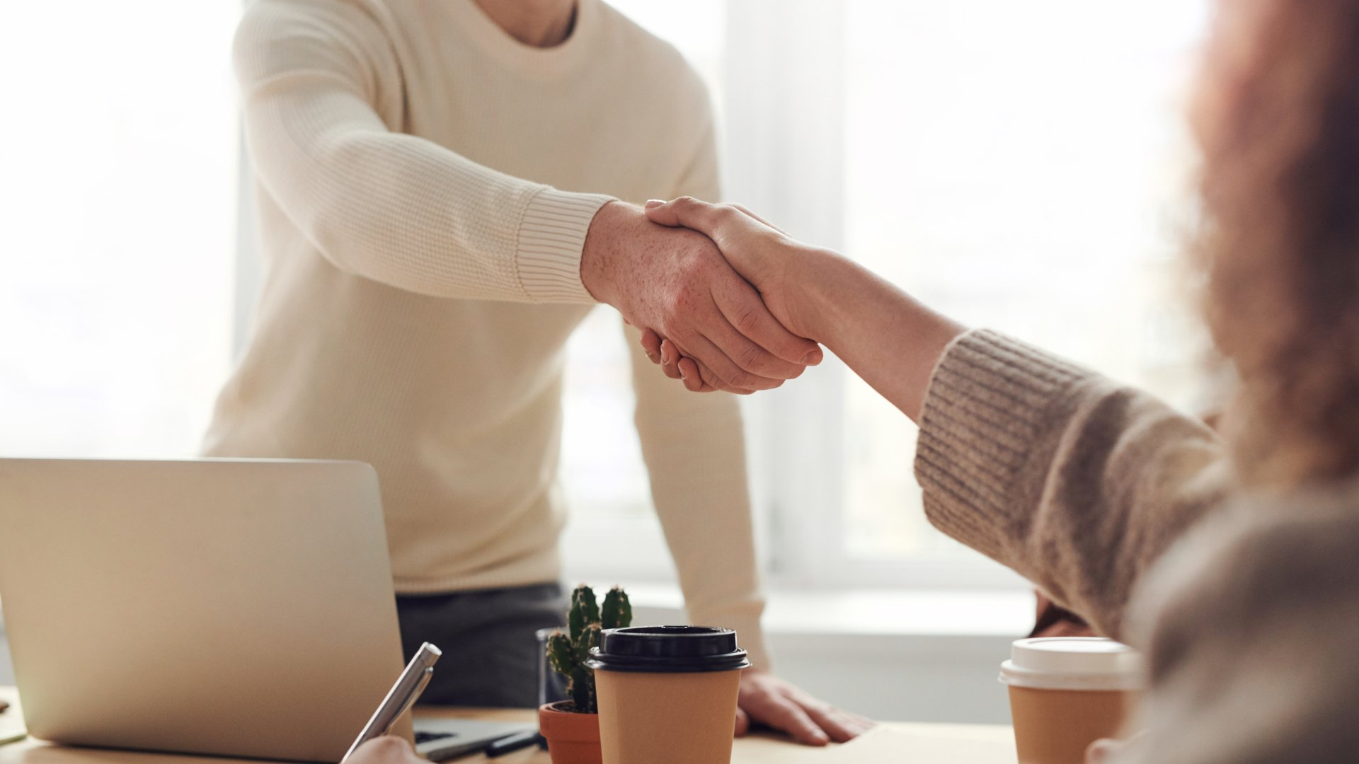Two people in neutral sweaters shake hands across a desk with laptops and coffee cups.