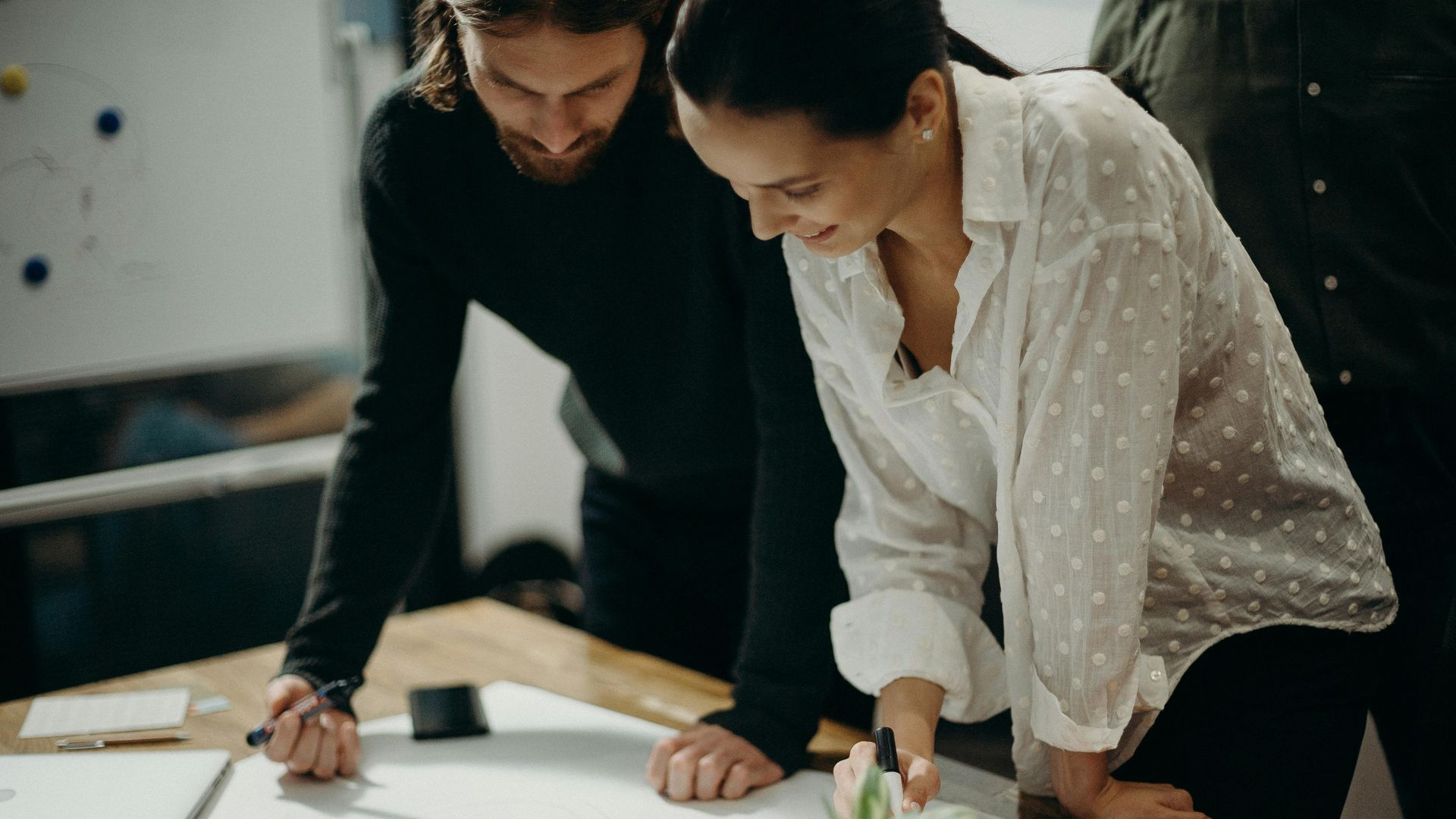 Two colleagues collaborate over papers on a wooden table in a brightly lit office setting.