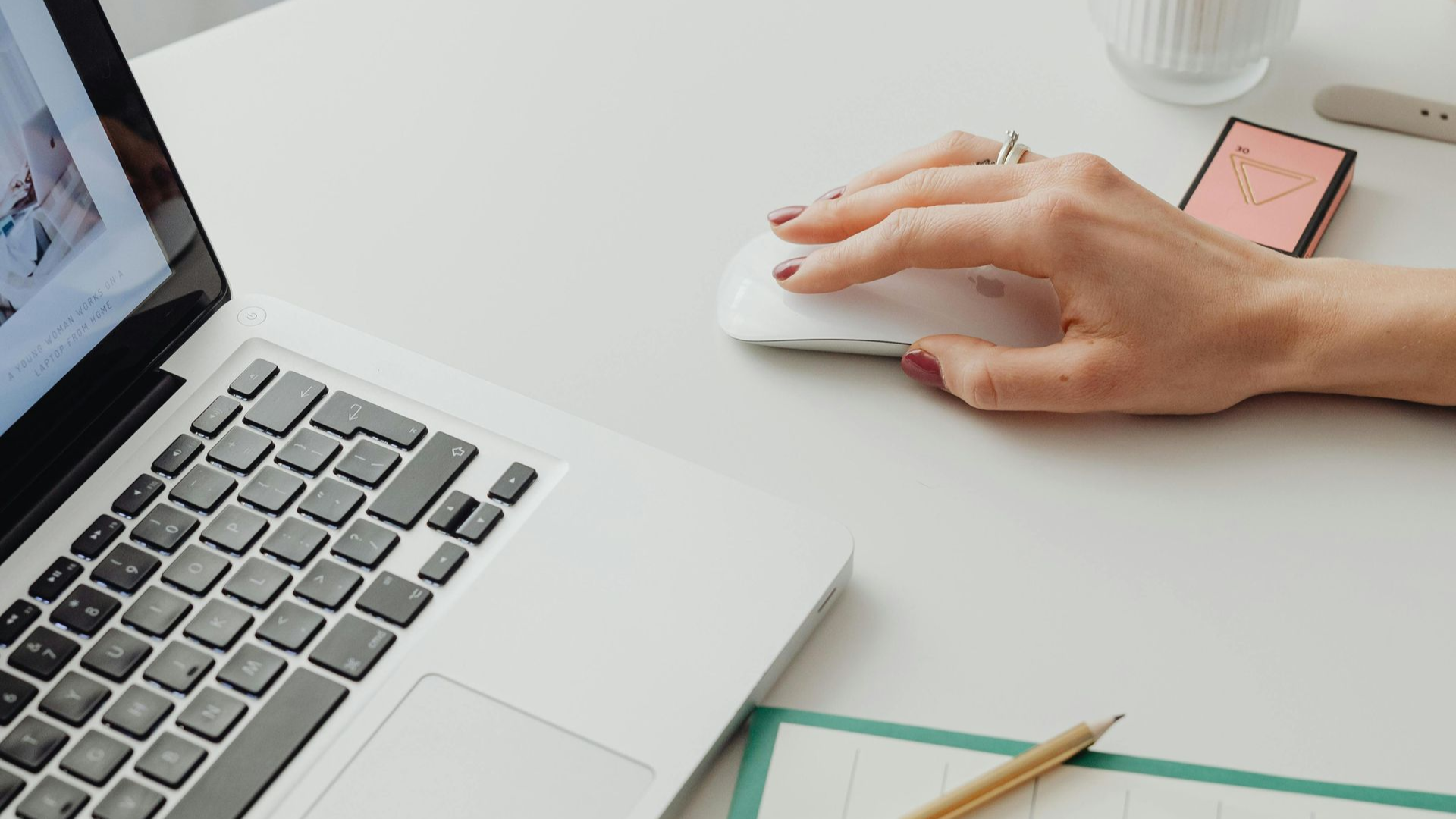 A person’s hand uses a white computer mouse on a white desk next to a laptop, a notebook, and a pencil.