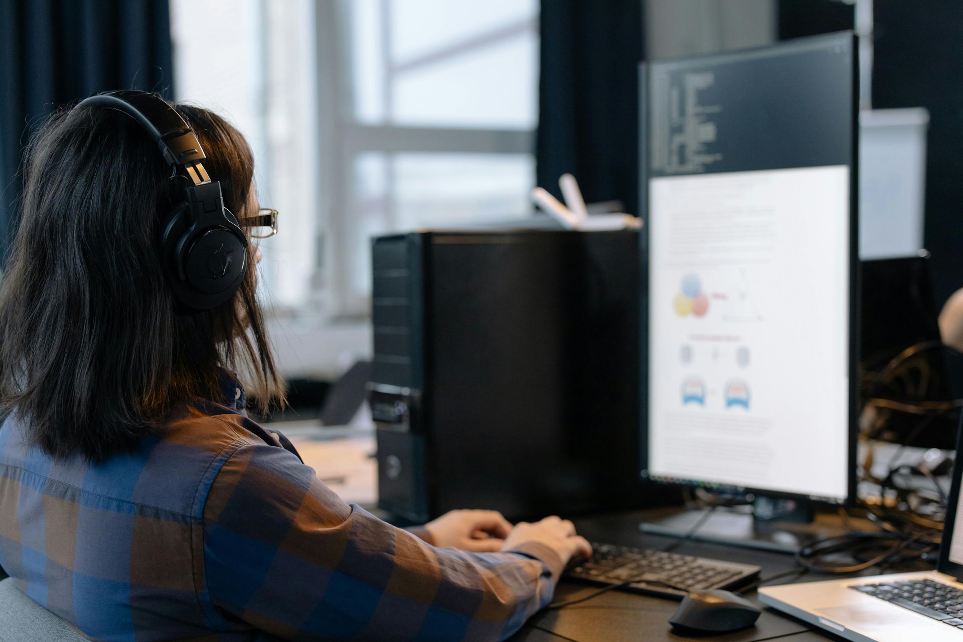 A person wearing headphones works at a computer in a workspace with a vertical monitor displaying diagrams.