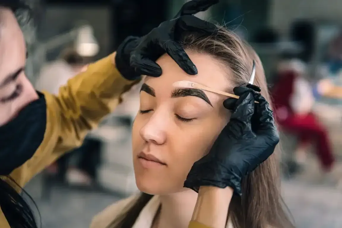 Eyebrow shaping: Person's brow being waxed. Technician in black gloves, mask, holds tools over client's closed eyes.
