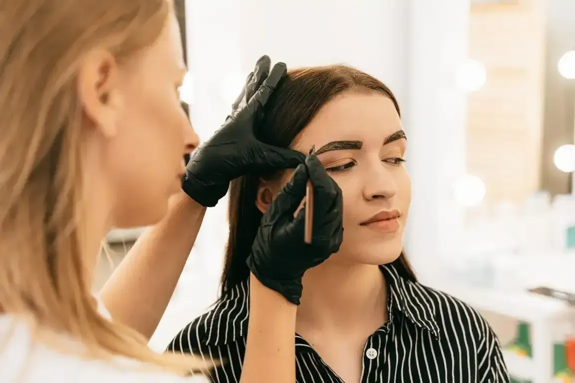 Woman receiving eyebrow tinting by master lash tech with black gloves.