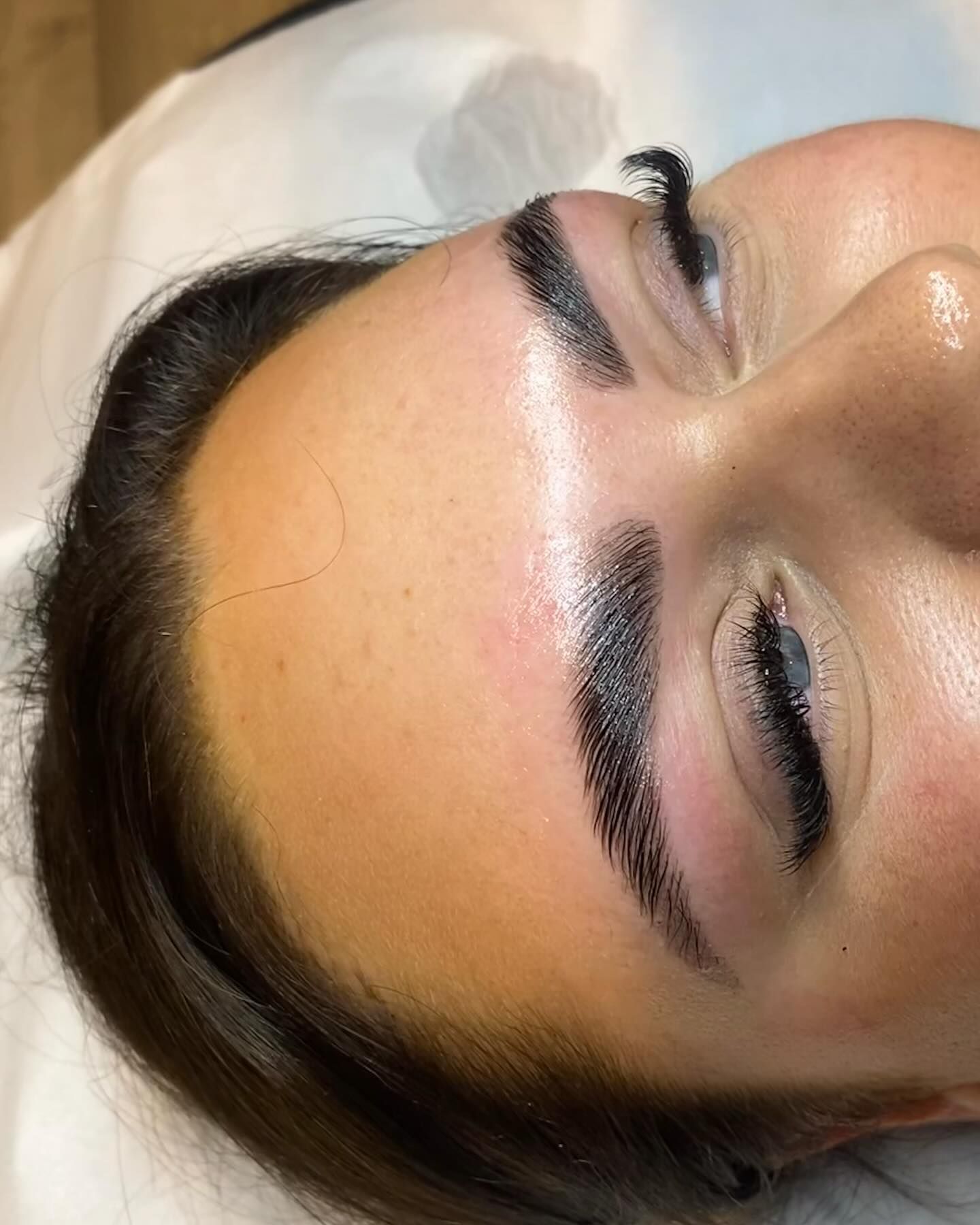 Close-up of a person with dark, groomed eyebrows, lying down. Their skin has a slight shine.