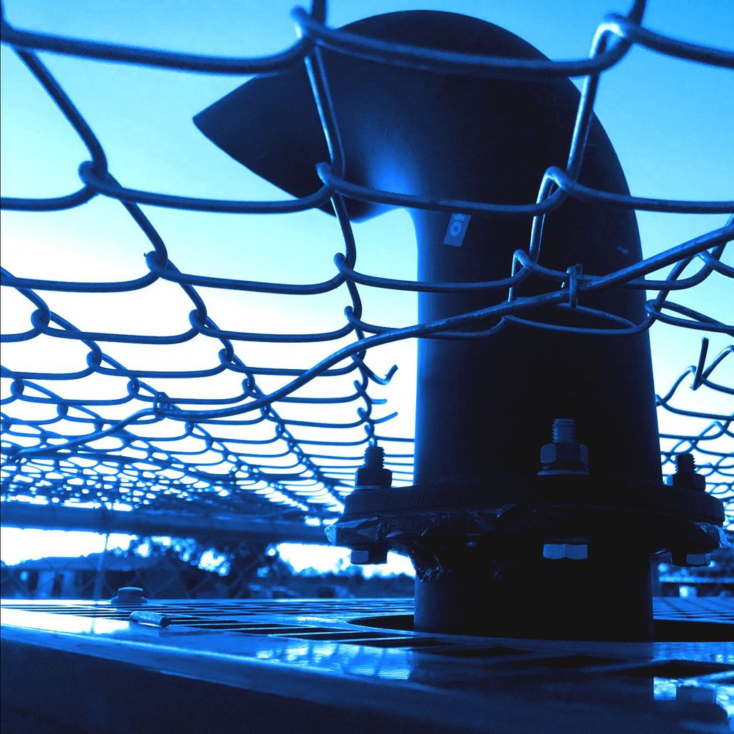 A black pipe is behind a chain link fence with a blue sky in the background