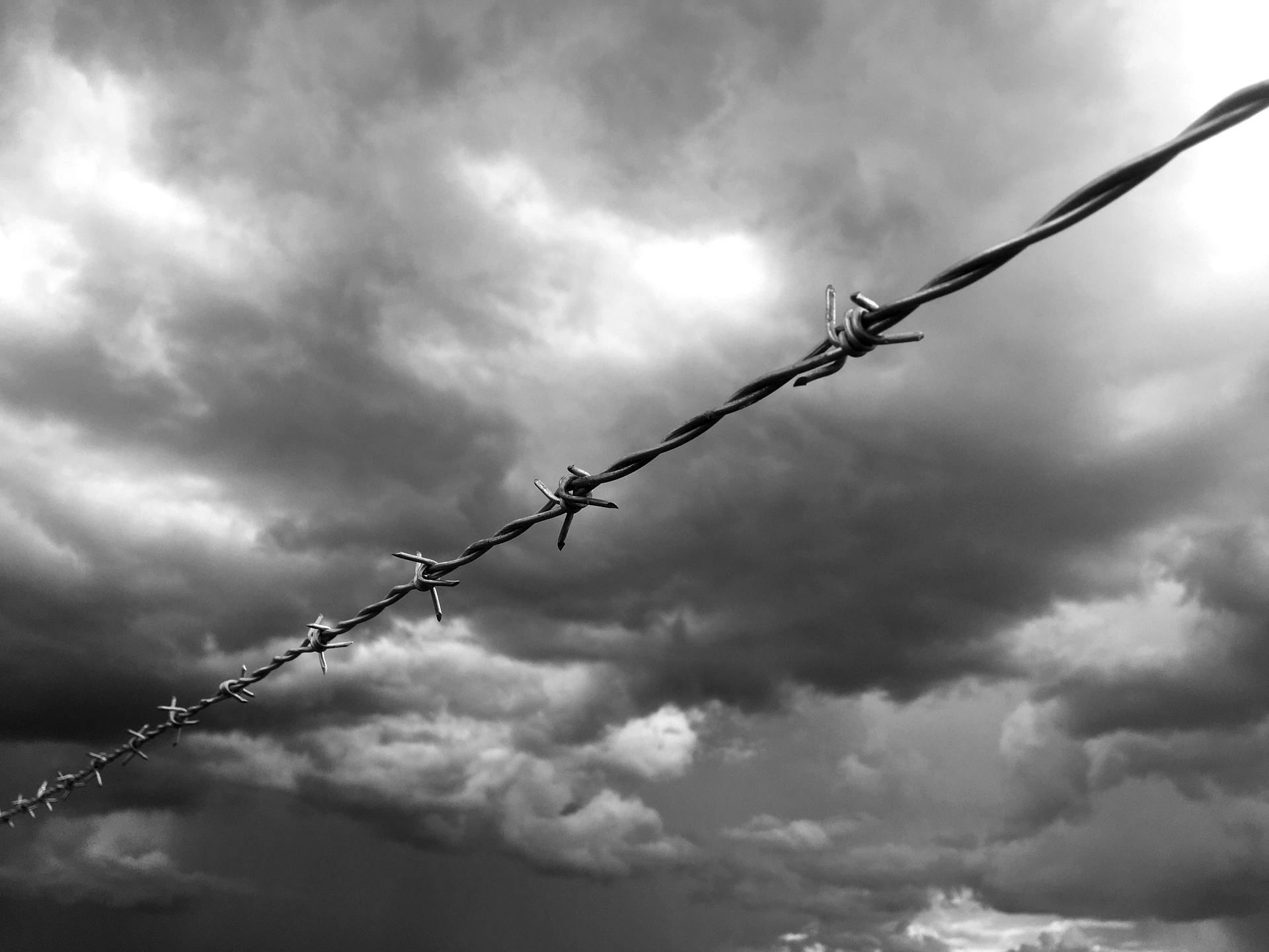 A black and white photo of a barbed wire fence against a cloudy sky.