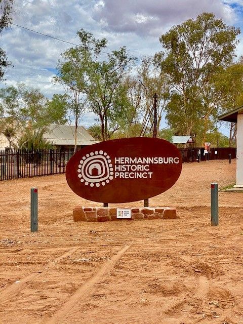 A sign for hermannsburg historic precinct is in the middle of a dirt field.