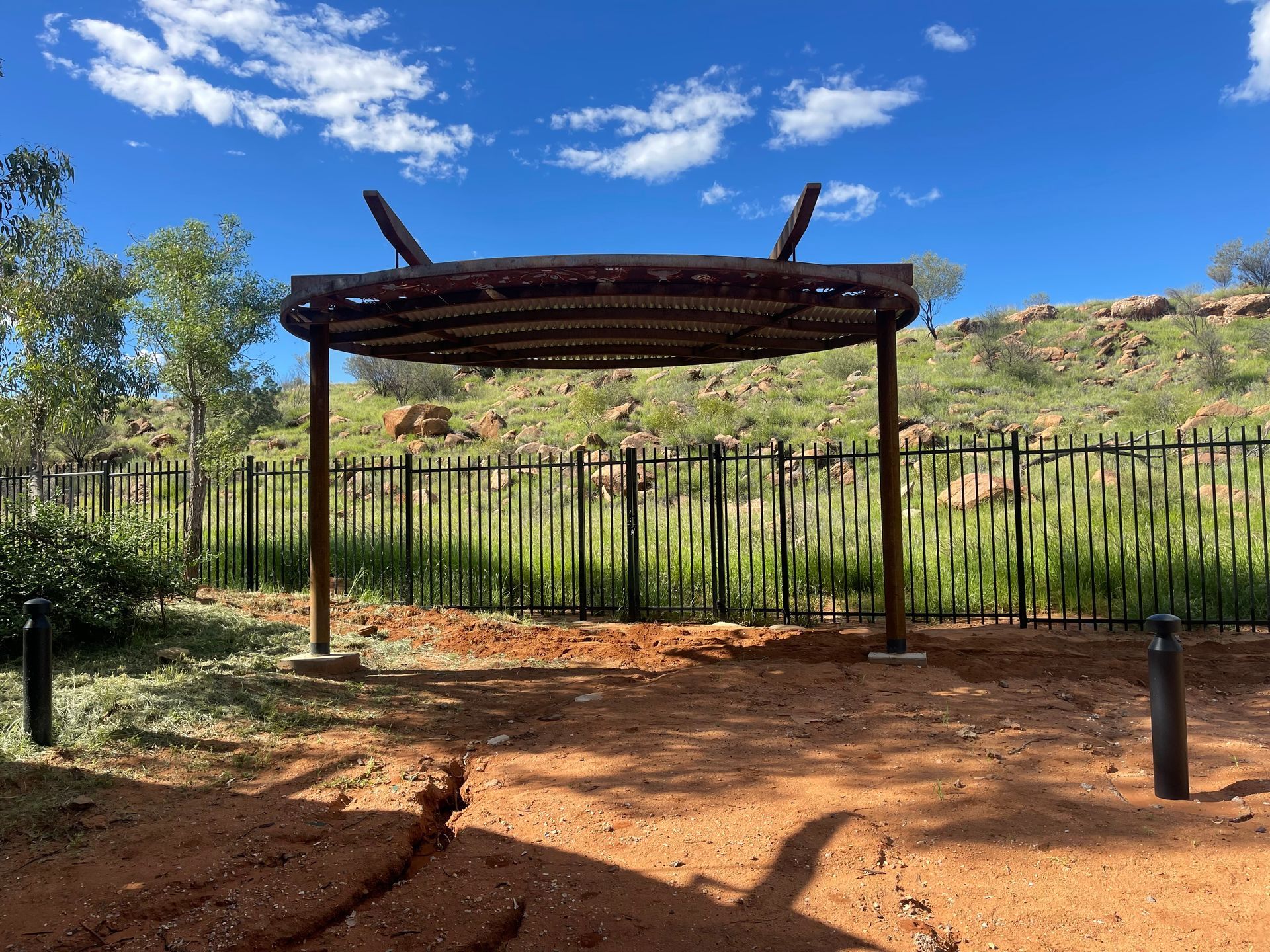 A wooden pergola is sitting in the middle of a dirt field next to a fence.