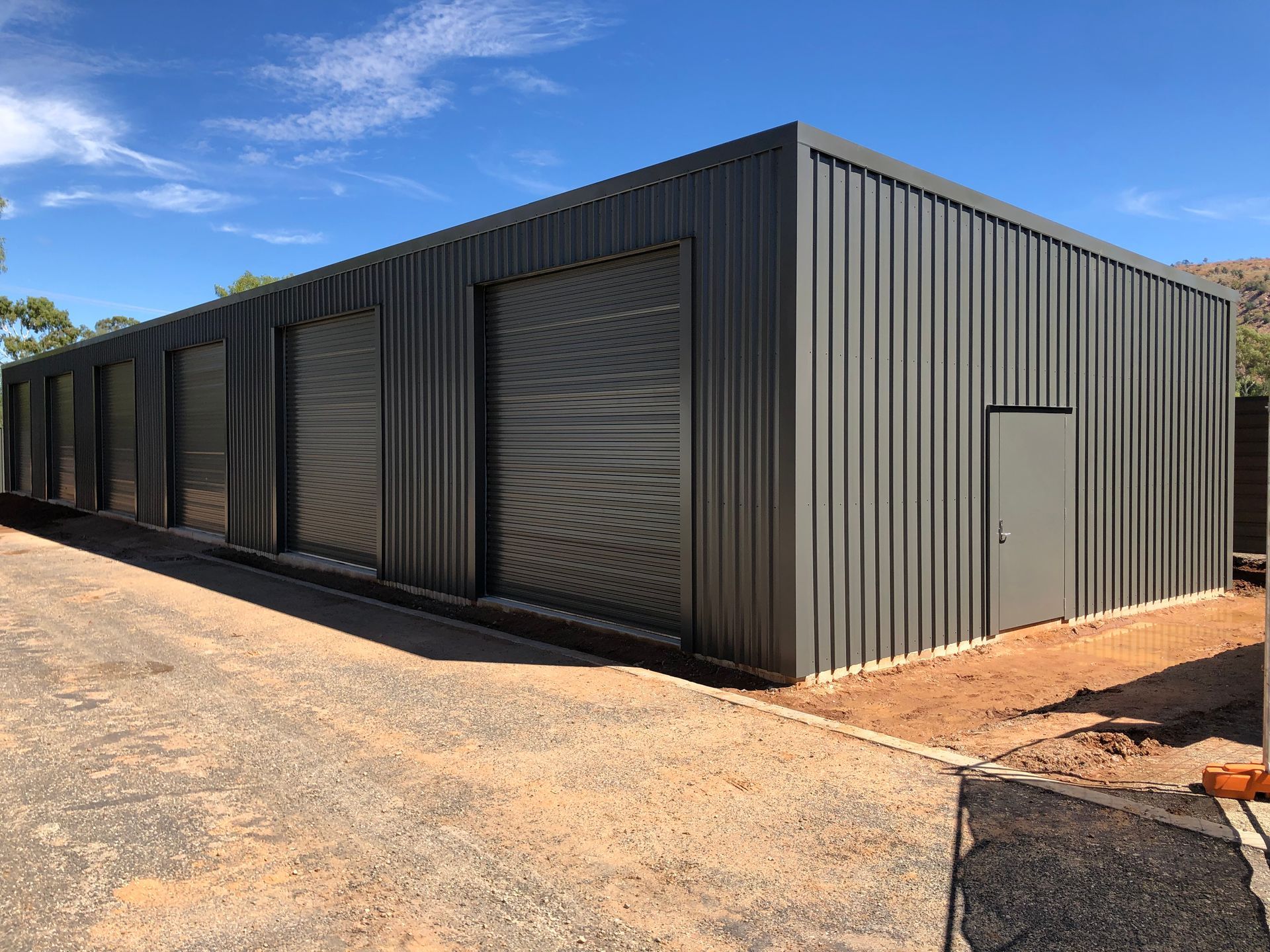 Gray metal building with roll-up doors and a side door on a gravel lot under a blue sky.