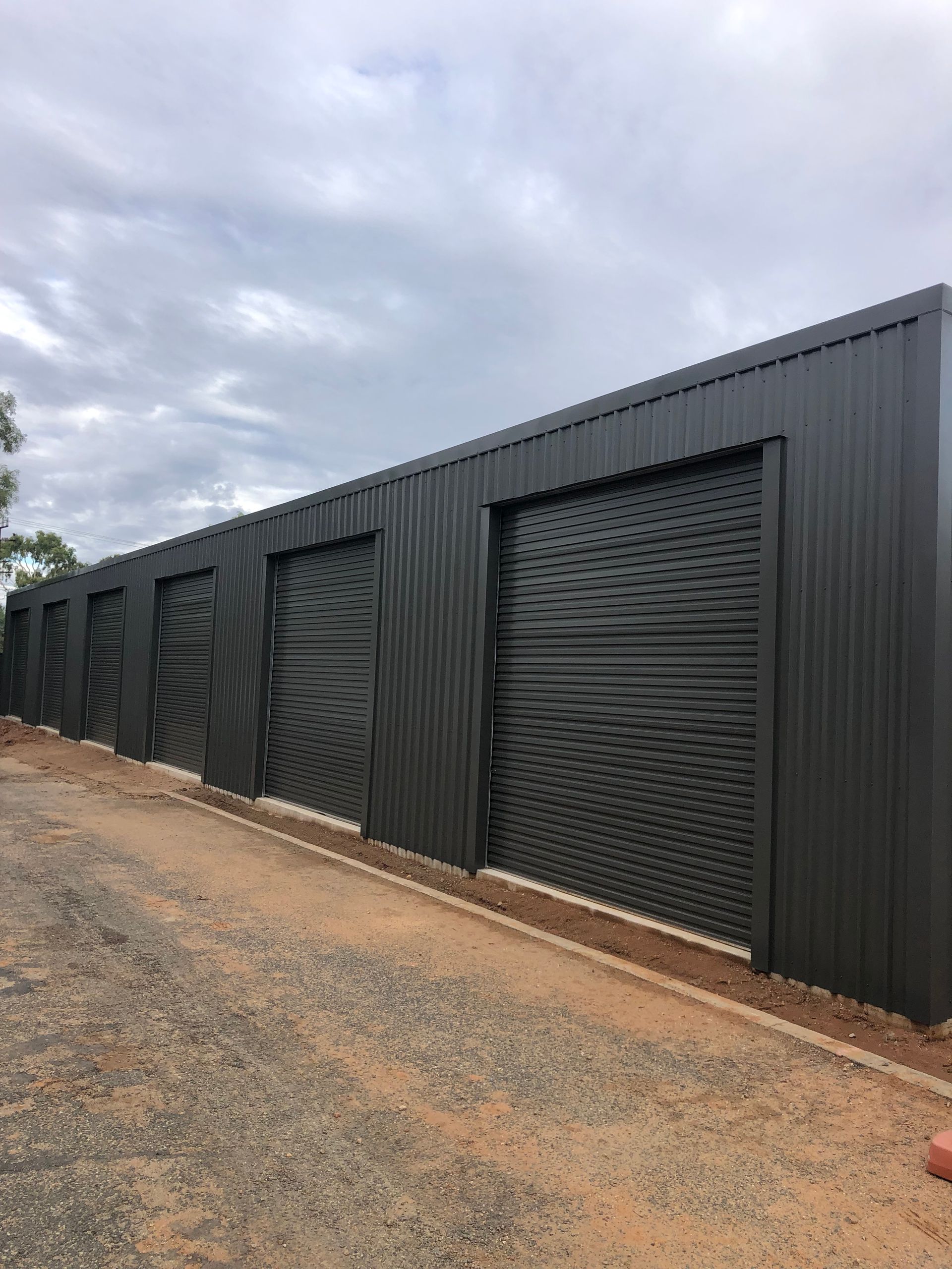 Black storage units with patterned doors, lined along a gravel driveway. Cloudy sky overhead.