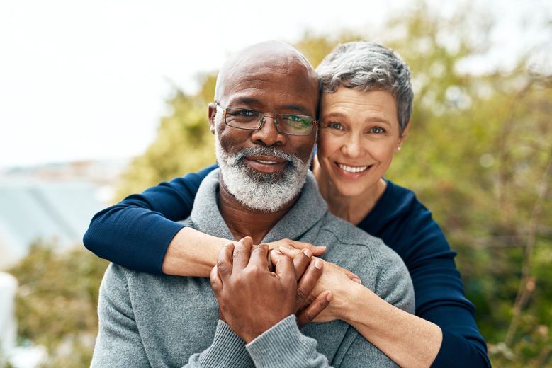 Couple embracing outdoors, woman's arms around man's shoulders, holding hands, smiling.