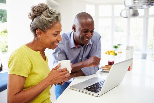 Couple smiles at laptop in bright kitchen. Woman holds a mug, man gestures.