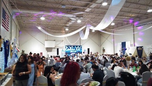 Large hall with a gathering; people seated at tables, decorated with lights and fabric.