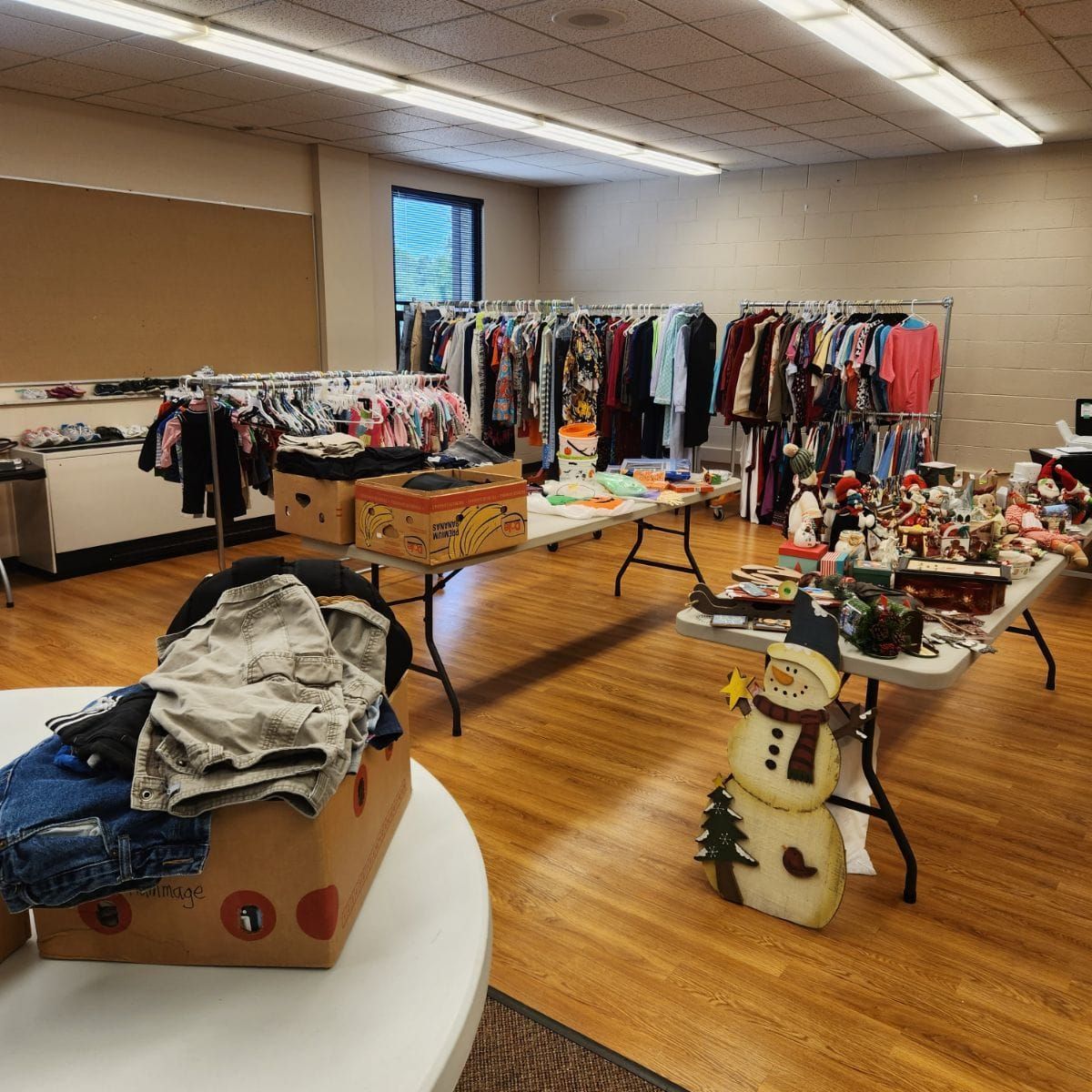 Room with clothes racks, tables displaying items, and a snowman decoration, likely a sale.