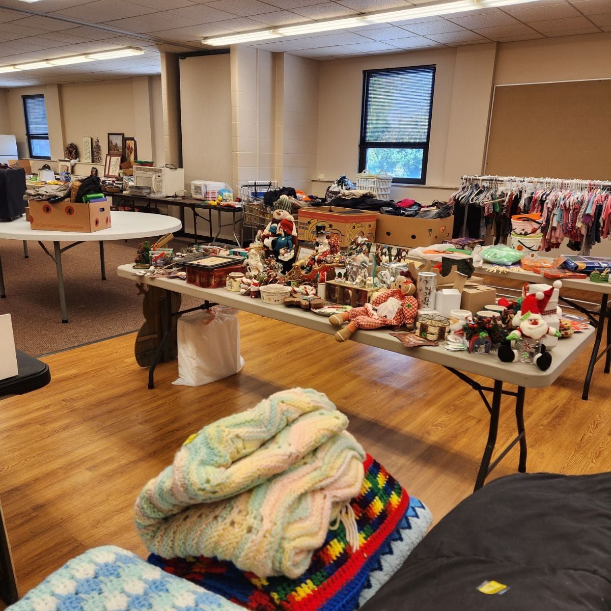 Room with tables laden with items, possibly a sale. Blankets, clothes, and various goods are visible.