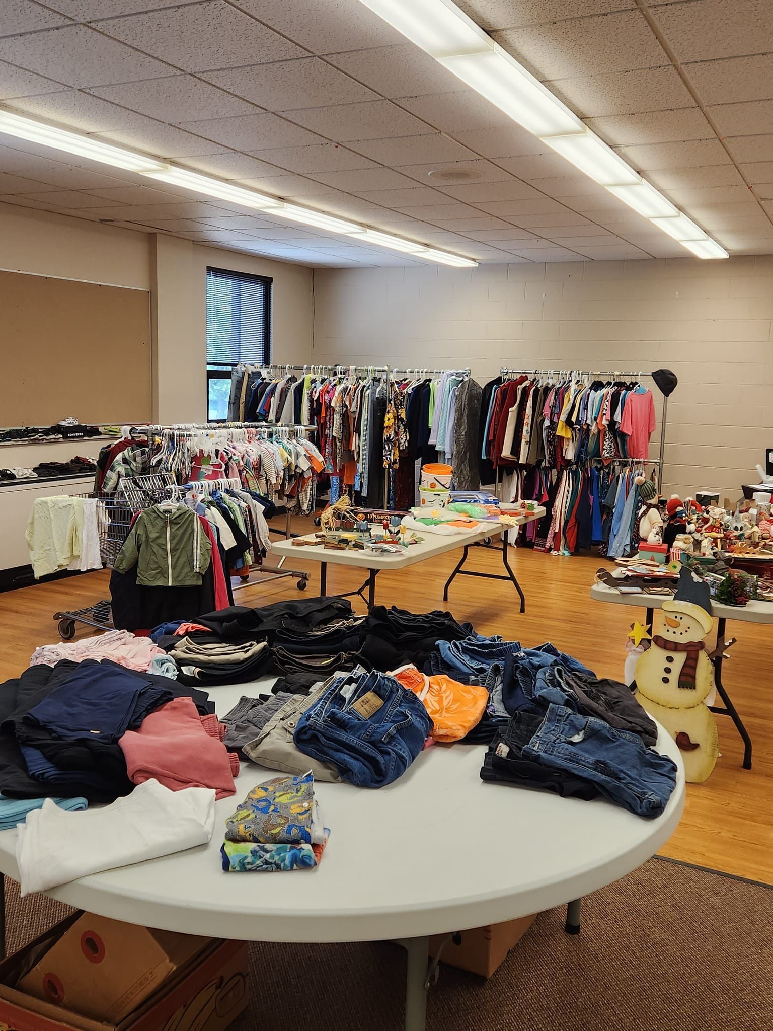 Clothes and items displayed on tables and racks at a community sale inside a brightly lit room.
