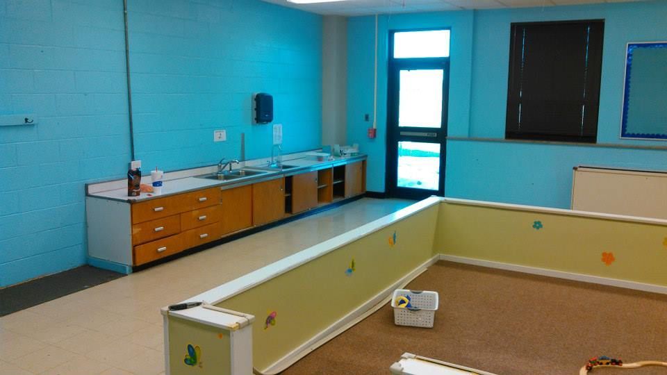 A light blue classroom with a sink, wooden cabinets, and a yellow play area with toys.