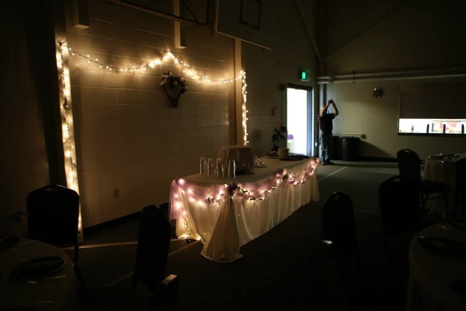 Wedding reception setup with string lights, table, and decorations in a dimly lit room.