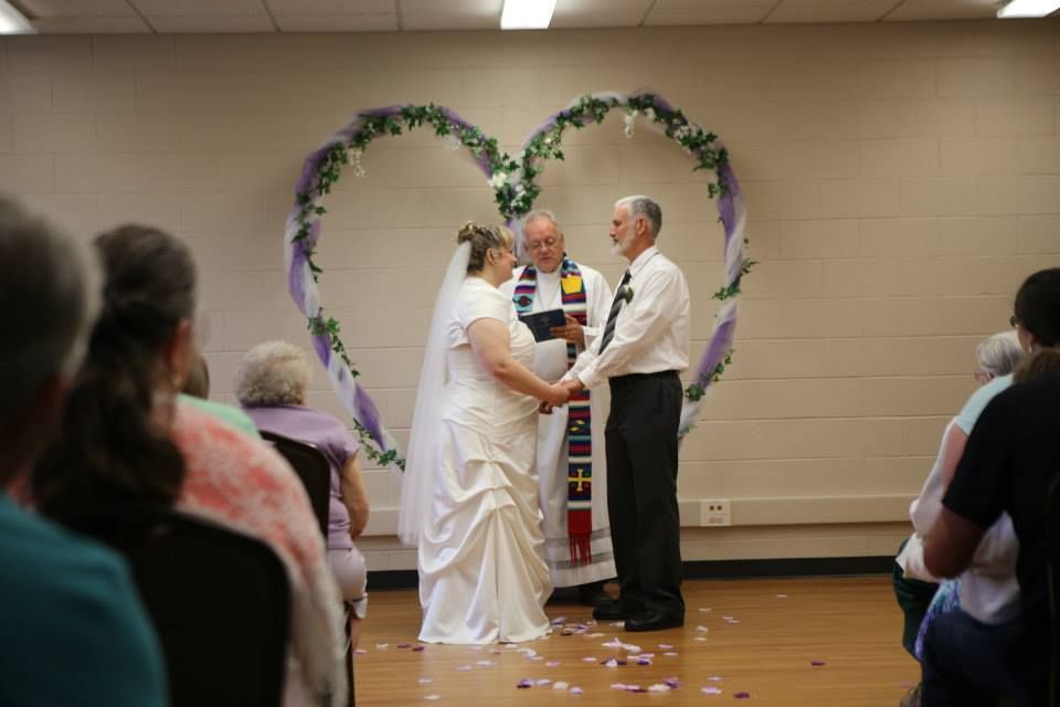 Couple exchanging vows during a wedding ceremony in front of a heart-shaped floral arch. Guests watch.