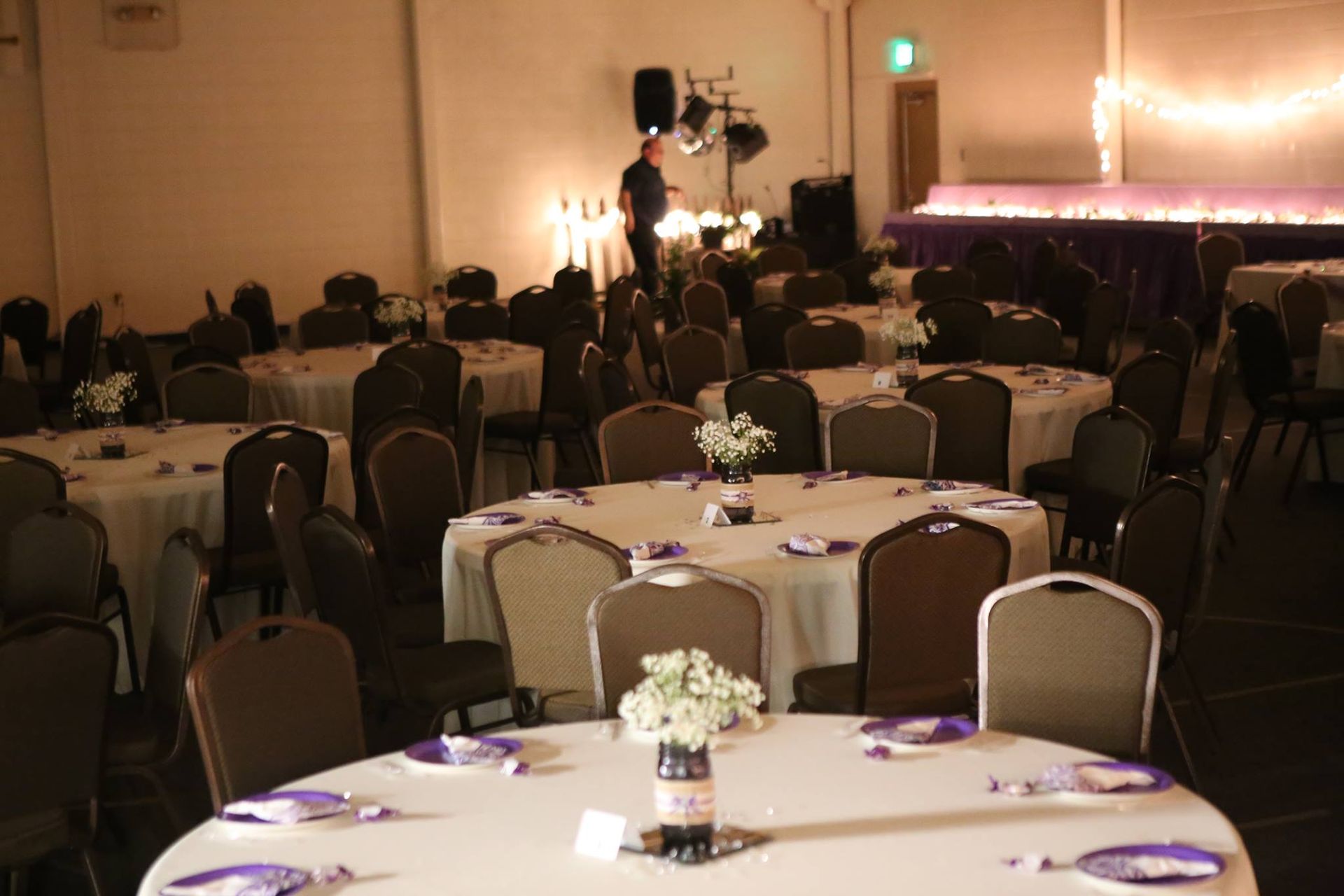 Banquet hall with round tables set for a formal event; person setting up lights in the background.
