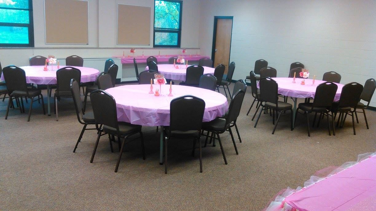 Round tables draped in pink, set with black chairs in a room with light carpet.