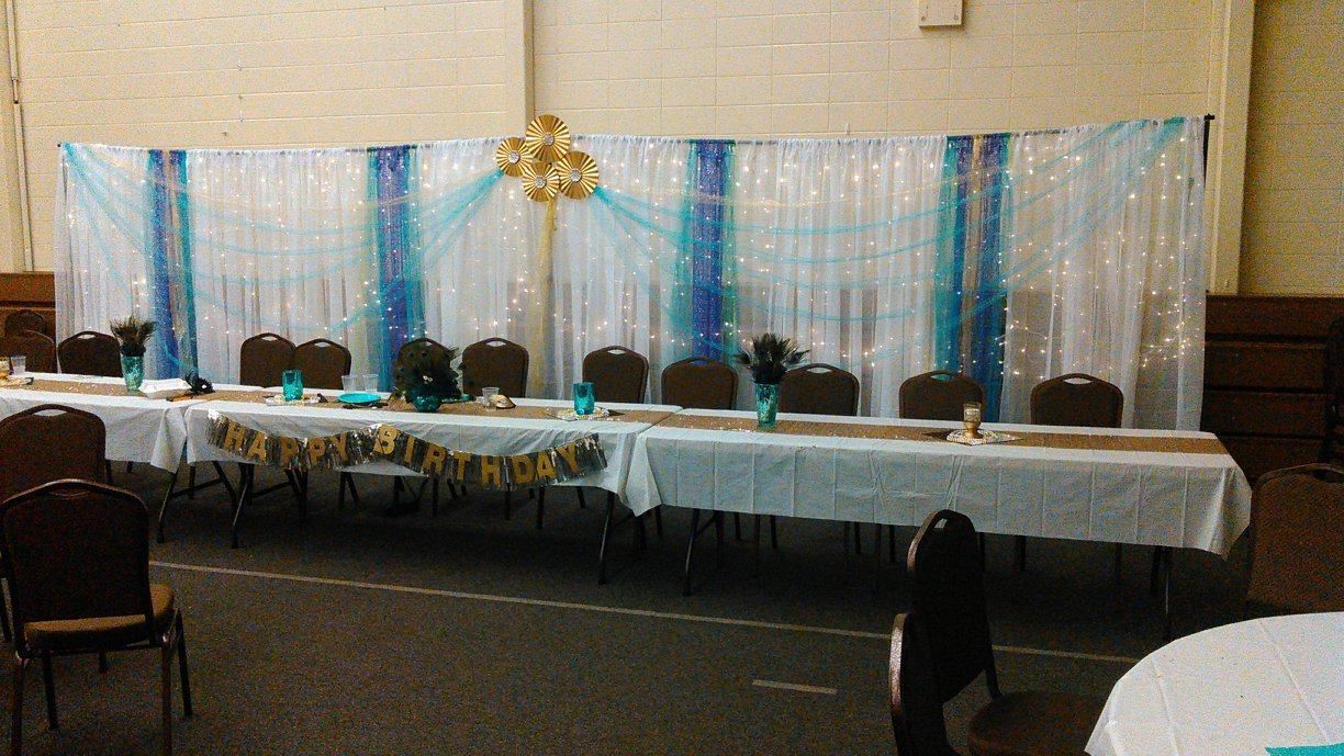 Long banquet table set for a special event with a decorated backdrop of white, turquoise, and gold.