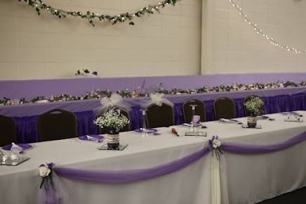 Long white table decorated with purple fabric, flowers, and bows at a wedding reception.