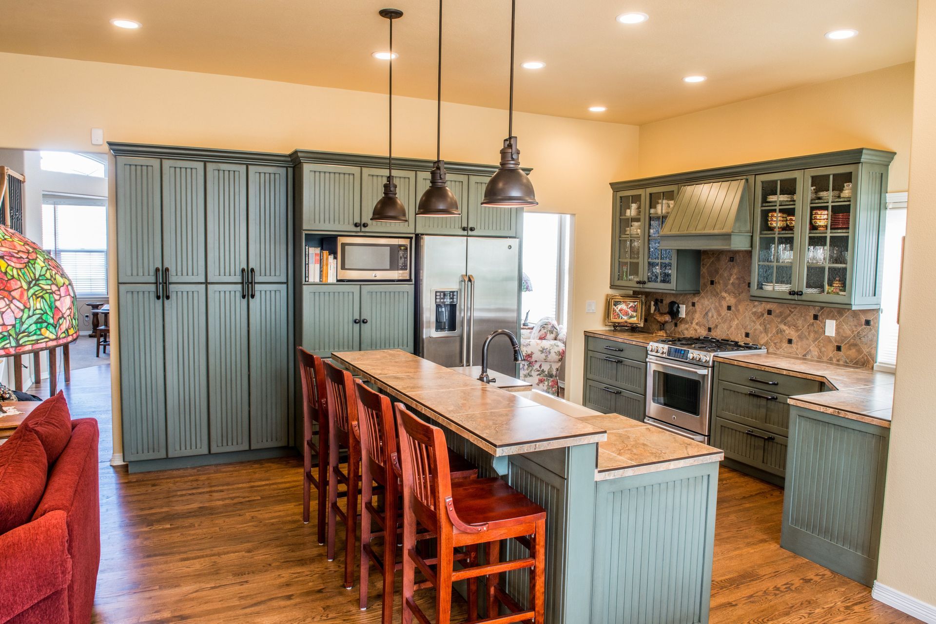 A kitchen with sage green cabinets, a wood-topped island with three barstools, stainless appliances, and wooden floors.