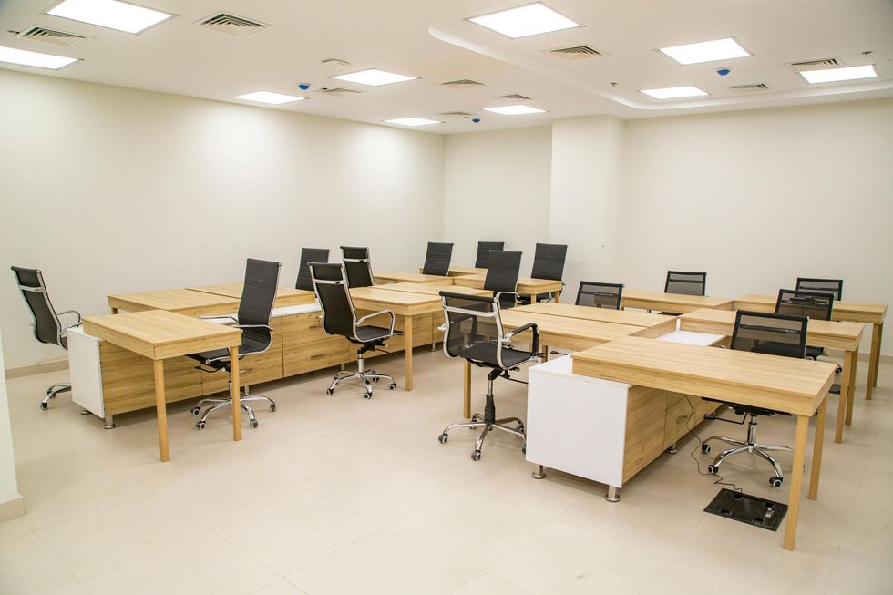 A bright, modern office space with multiple wooden desks and black ergonomic chairs arranged in rows.