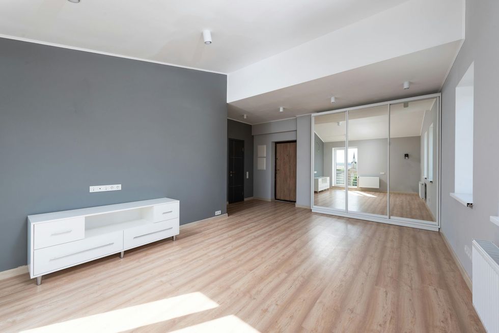 A modern, unfurnished room with light wood floors, a gray accent wall, a white media console, and a large mirrored closet.