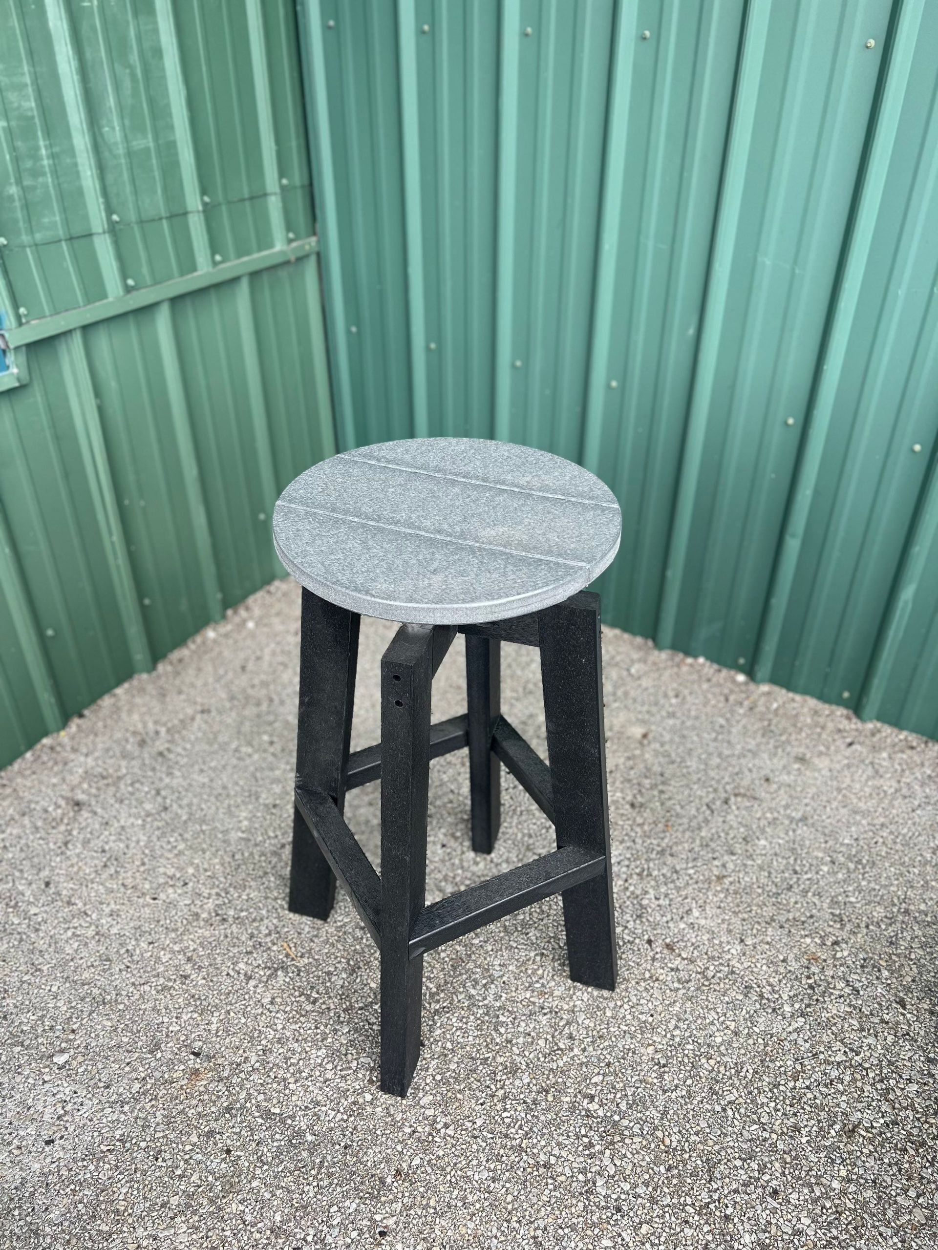 A black stool with a gray top is sitting on a gravel floor in front of a green fence.