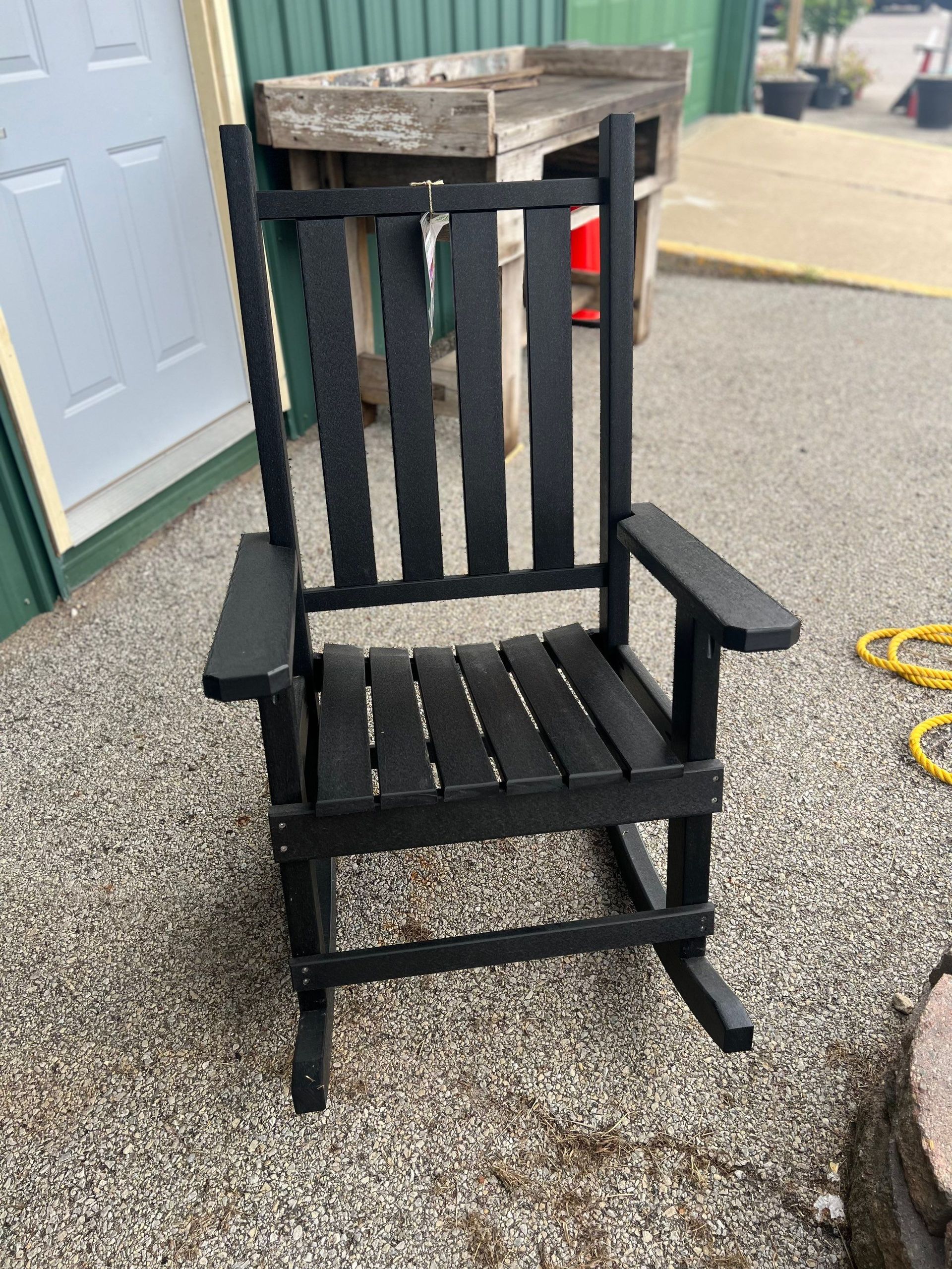 A black rocking chair is sitting on gravel next to a hose.