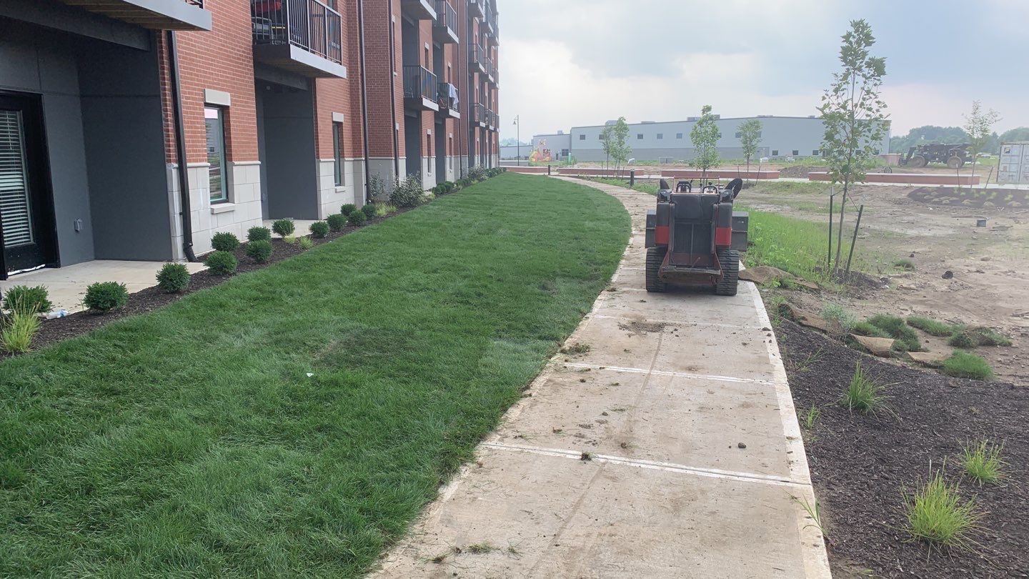 A lawn mower is driving down a sidewalk next to a building.
