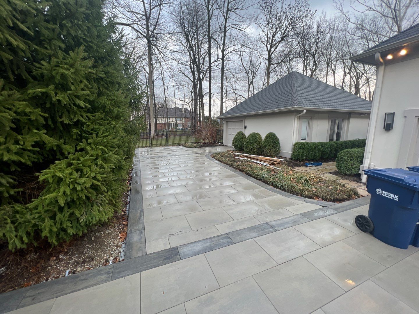 A blue trash can is sitting on the side of a driveway next to a house.