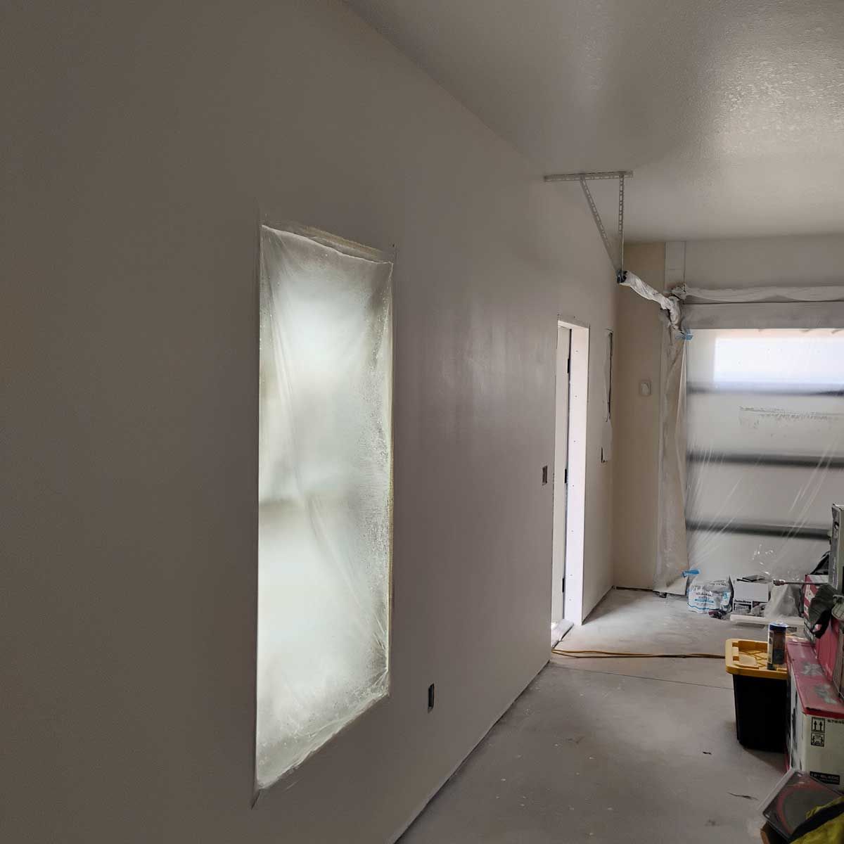 Interior view of a room under renovation. White walls with a covered window, door, and unfinished garage door are visible.