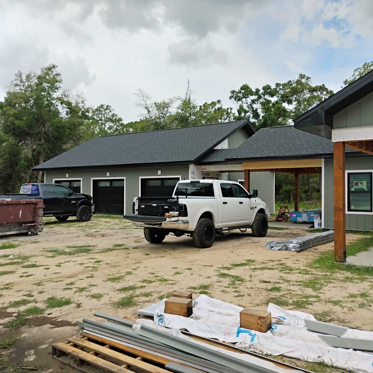 Construction site with a white pickup truck in front of a gray house with black garage doors and a green house to the right.