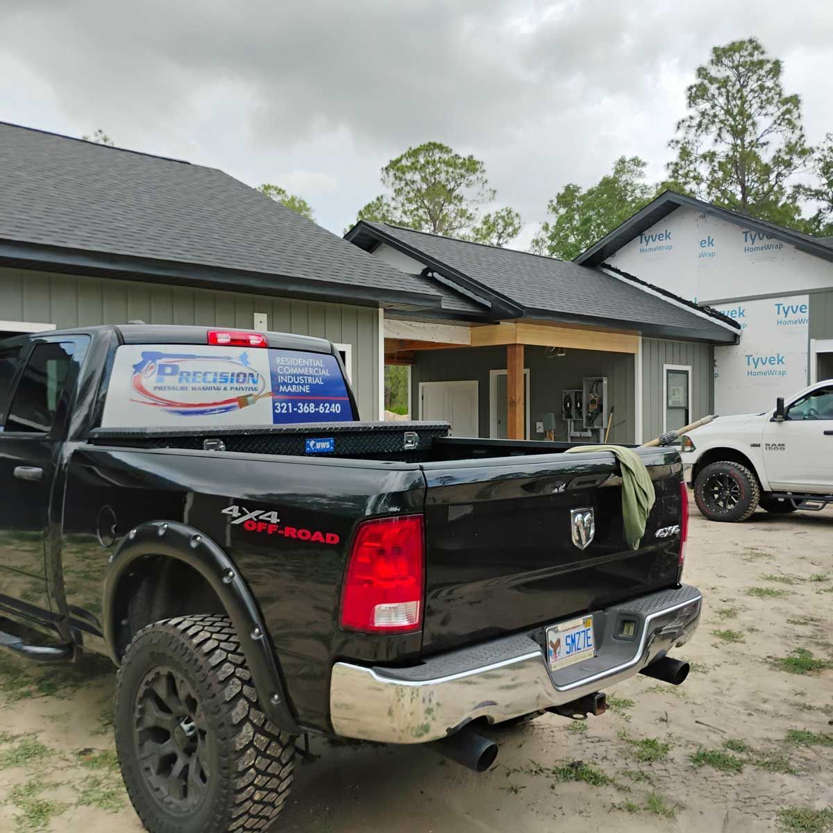 Black pickup truck parked in front of a house under construction; a white truck is visible too. The truck has a company logo.