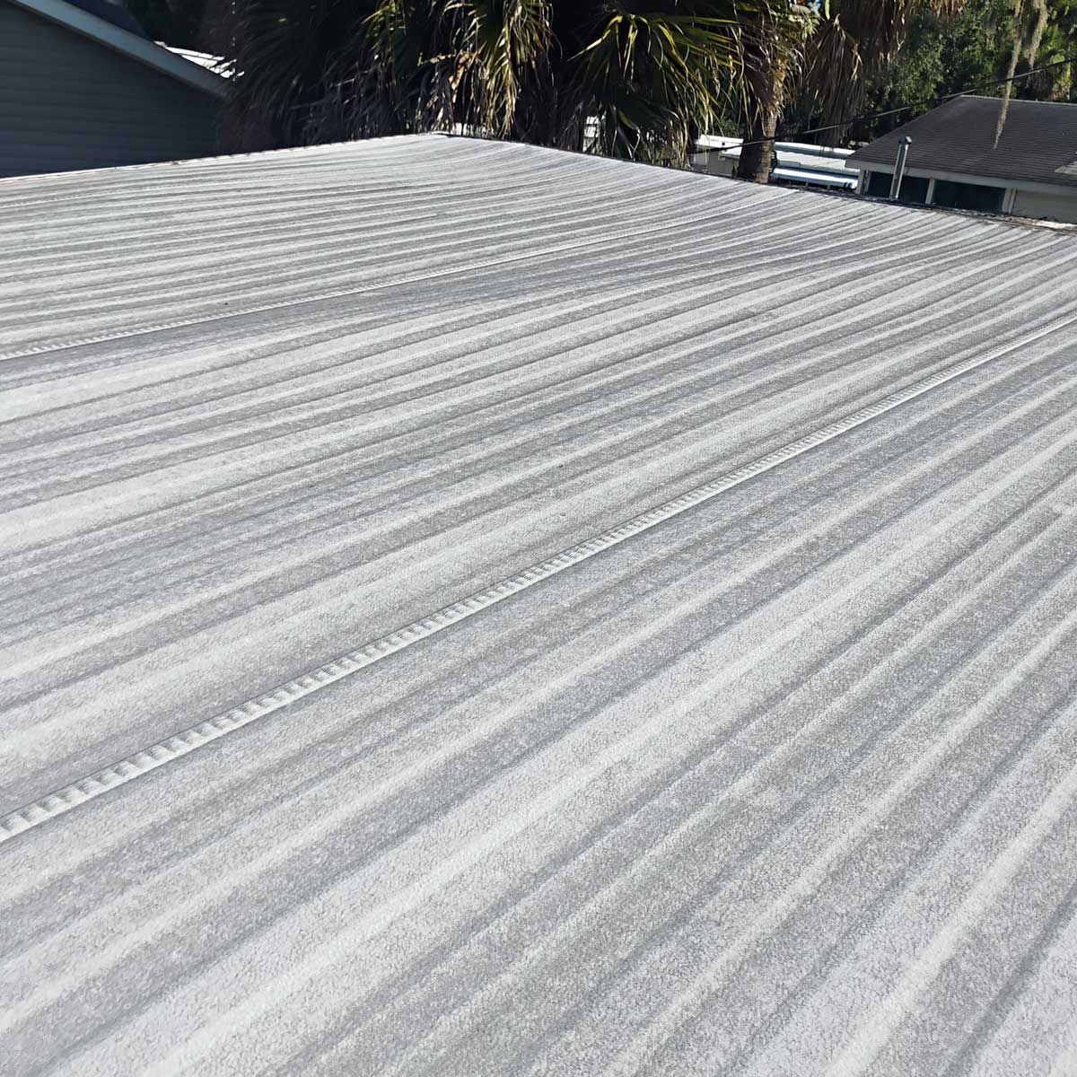 Close-up view of a gray, corrugated metal roof under bright sunlight, with palm trees in the background.