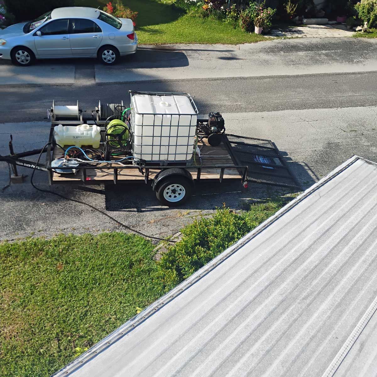 A trailer with pressure washing equipment parked on a residential street, with a silver car and roof visible.