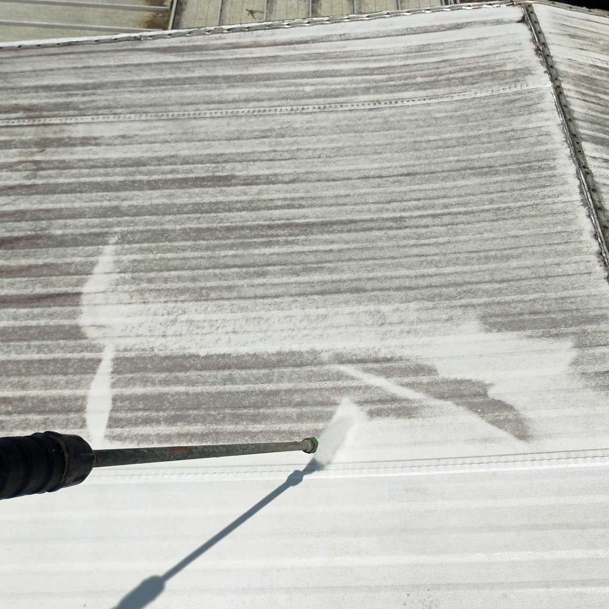 A person pressure washing a light-colored, ridged roof, leaving a bright clean streak.