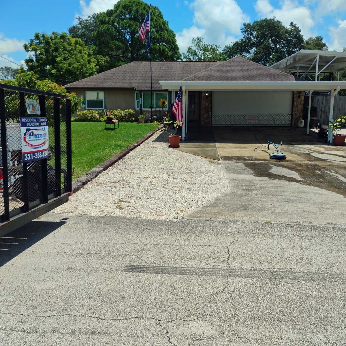 House with a driveway and a gravel area. An American flag flies in front. A gate is on the left.