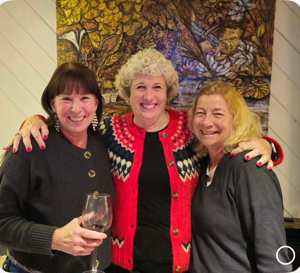 Three women smiling with their arms around each other in front of a patterned wall hanging; one holds a glass of wine.