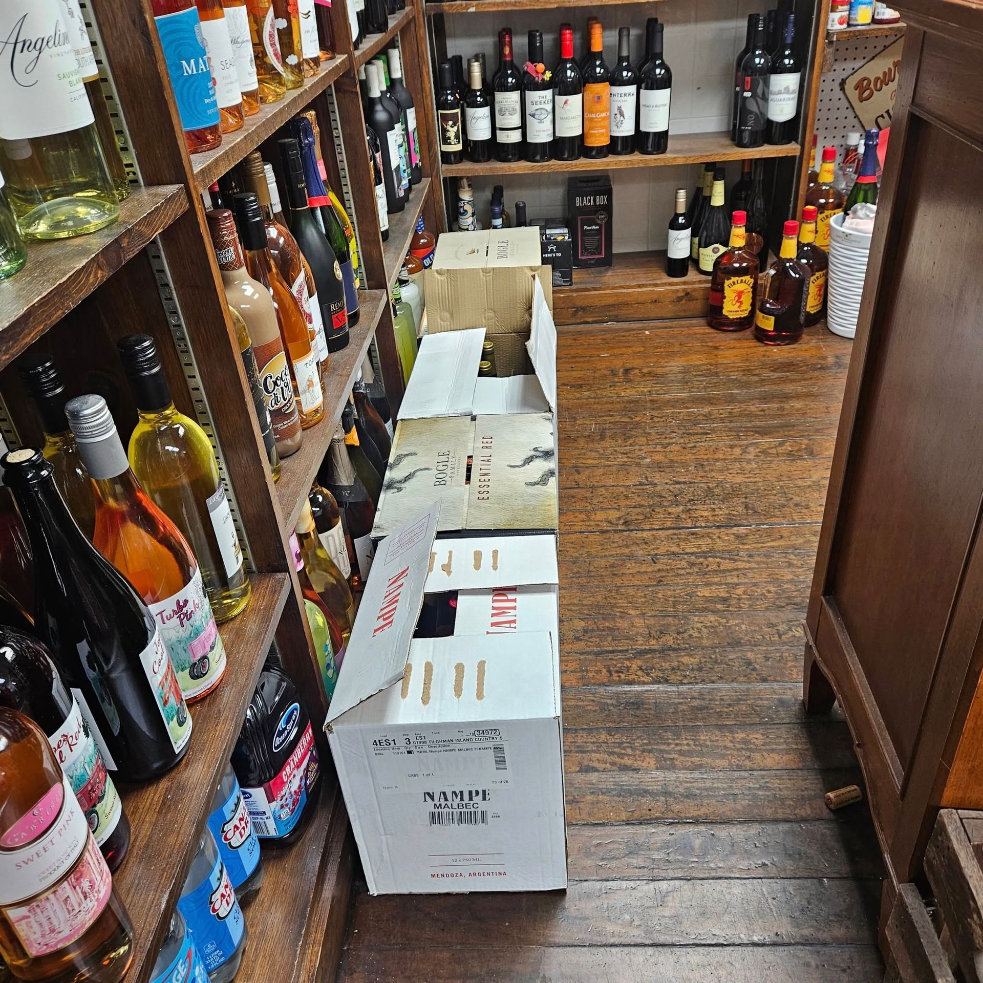 Shelves packed with wine and liquor bottles, with a row of cardboard cases sitting on the floor of a store.