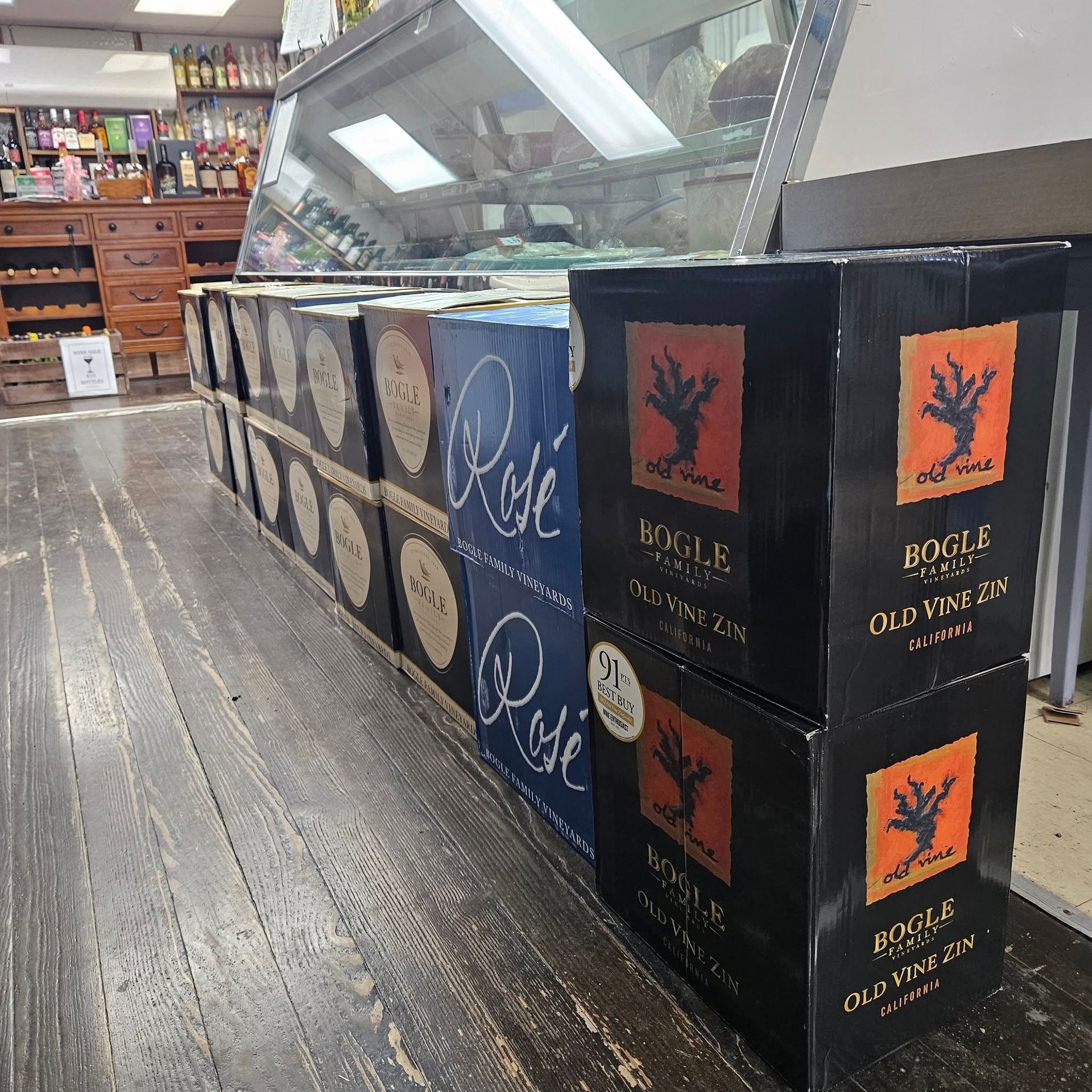 Rows of stacked wine boxes, including Bogle and Rosé brands, on a wood-plank floor inside a retail store.