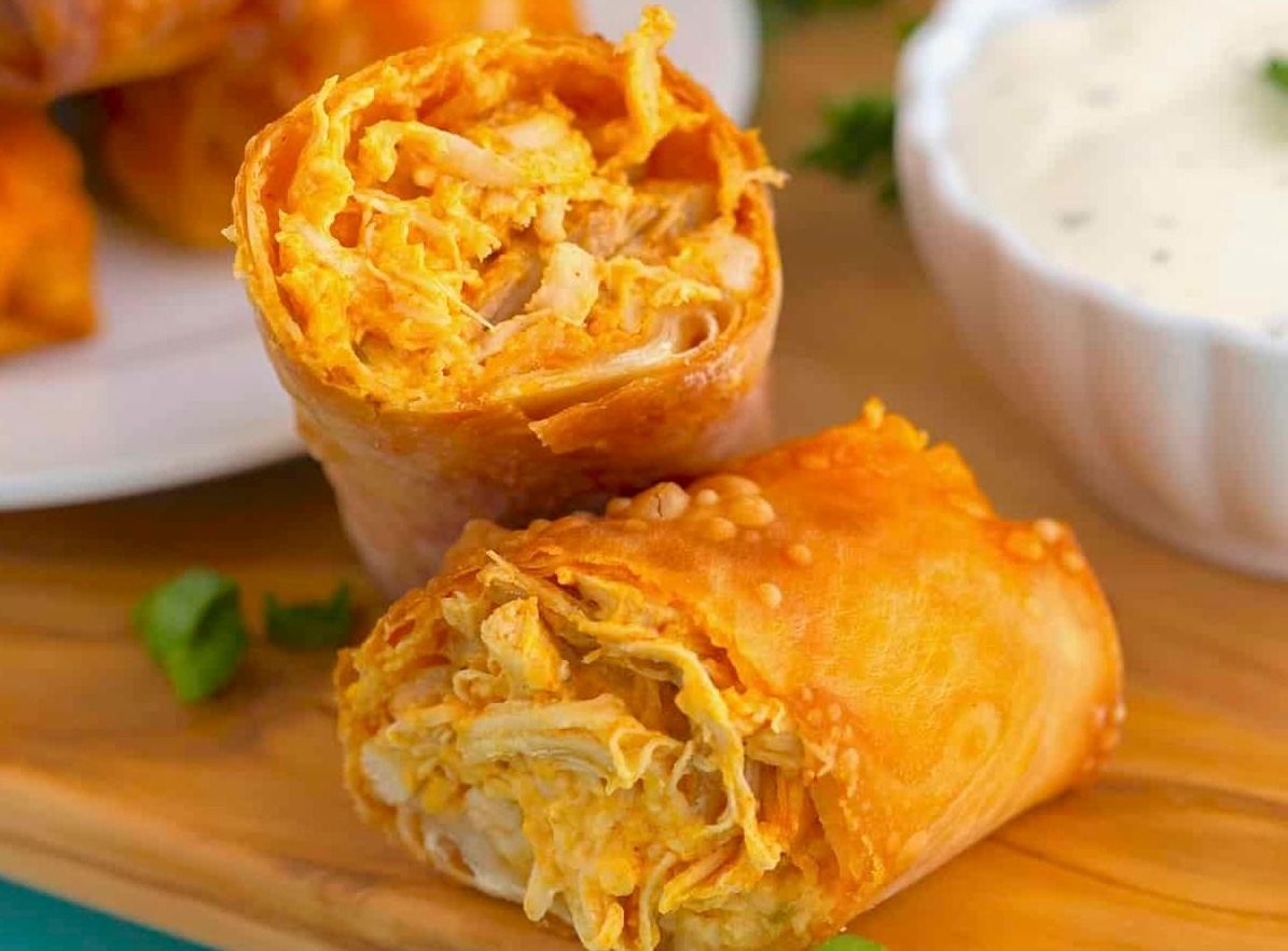 Two golden-brown crispy buffalo chicken egg rolls on a wooden board next to a small bowl of white dipping sauce.
