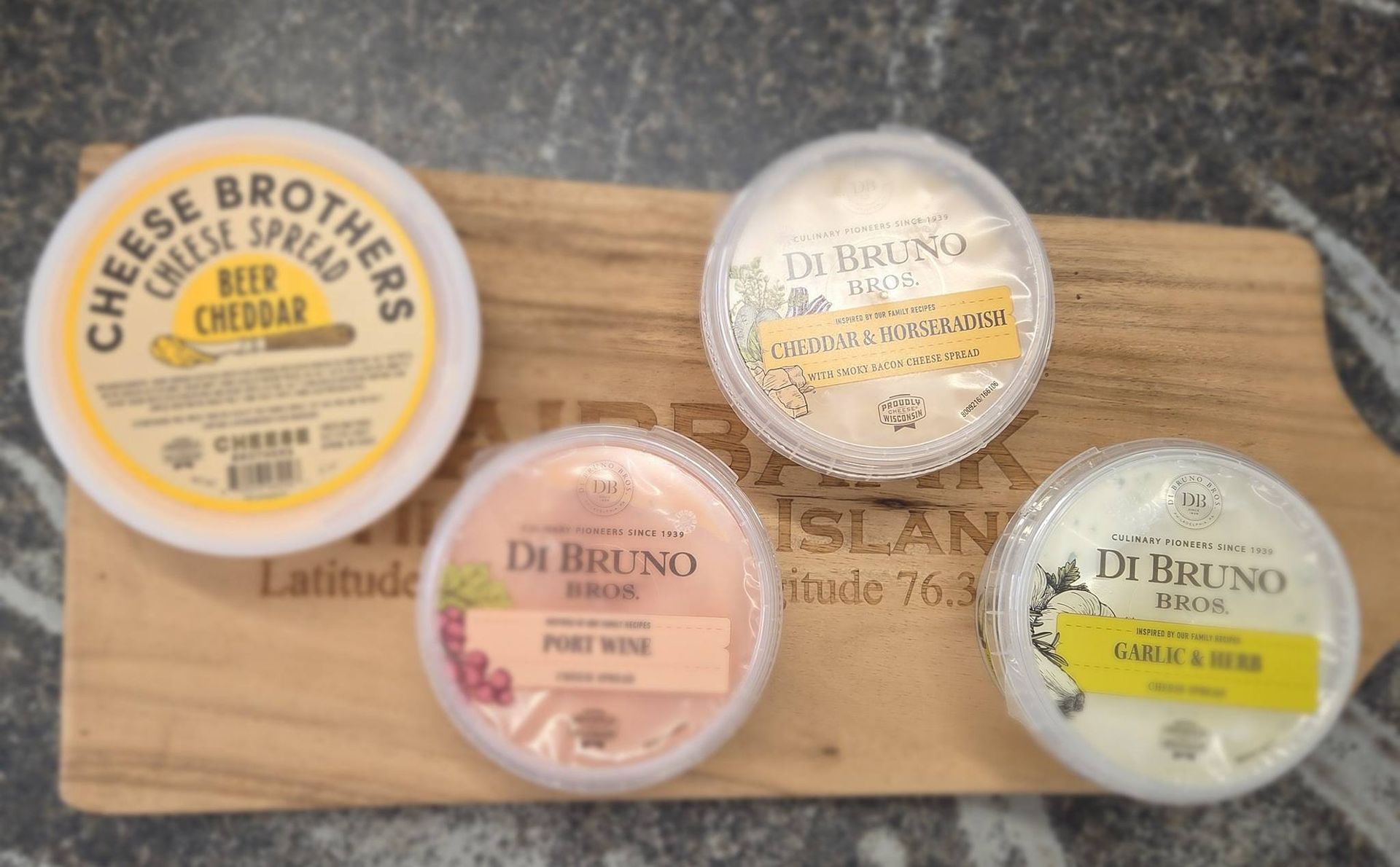 Four containers of cheese spread arranged on a wooden serving board, including one Beer Cheddar and three Di Bruno Bros.